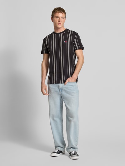 Männermodel trägt Regular Fit T-Shirt aus reiner Baumwolle von Tommy Jeans mit Optik:Allover-Muster, Label-Stitching-Applikation, Rundhals, Ärmelform: Kurzarm, Passform: Regular-Fit, in Schwarz mit weißen Streifen; glatte Textur; Ganzkörperansicht von vorne.