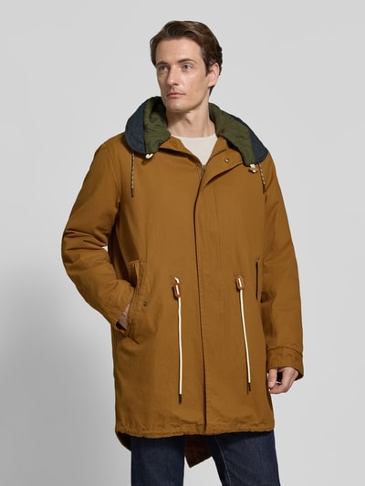 Männermodel trägt Regular Fit Mantel aus reiner Baumwolle Modell 'FISHTAIL' von Scotch & Soda in Braun mit Kapuze, Eingrifftaschen, Ganzkörperansicht von vorne.
