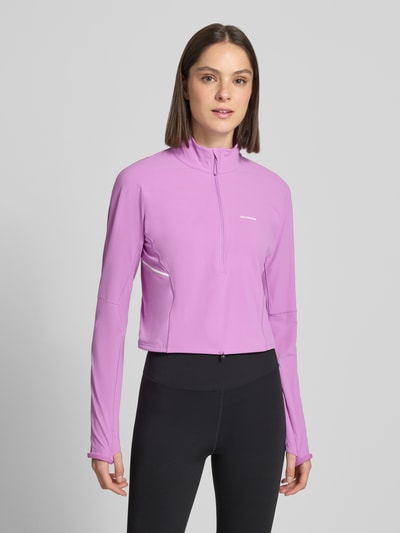 Modelka nosi bluzkę z długim rękawem 'Langarmshirt im Cropped-Design mit Stehkragen' od Salomon w taliowanym, krótkim kroju, stójka z ochroną podbródka, half-zip, tunel w pasie, kolor liliowy, długość tyłu w rozmiarze S: 47 cm, z długimi rękawami, całkowity widok z przodu.