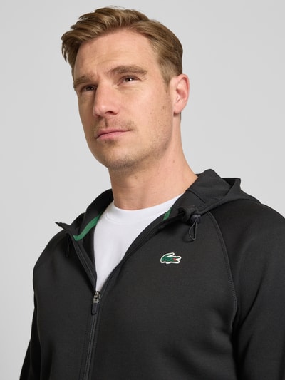 Mężczyznamodel nosi Regular Fit Sweatjacke mit Baumwoll-Anteil od Lacoste, efekt: jednolity kolor, krój regular fit, zamek błyskawiczny, naszywka z logo, kaptur z tunel, długi rękaw, boczne wpuszczane kieszenie, wykończenie materiału: scuba, mieszanka bawełny, zbliżenie na górną część ciała.