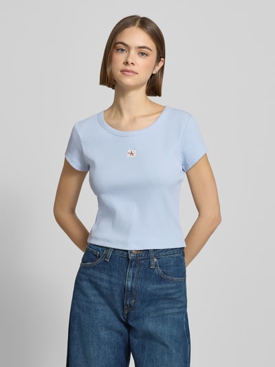 Kobiece modelka nosi Cropped T-Shirt mit Label-Detail od Calvin Klein Jeans z mieszanki bawełny i elastanu, okrągły dekolt, krój krótki, długość tyłu 43 cm dla rozmiaru S, jasnoniebieski kolor, gładka tekstura, widok całej sylwetki z przodu.