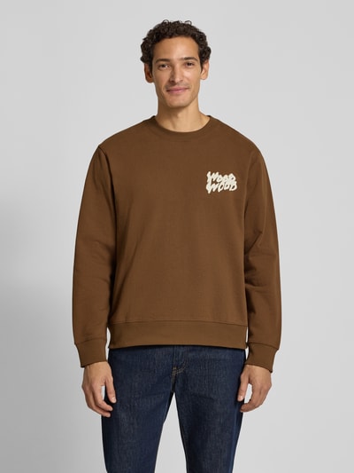 Männermodel trägt braunes Sweatshirt mit Label-Print Modell 'Colter' von Wood Wood, Rundhalsausschnitt, Regular Fit, Ganzkörperansicht von vorne. Reine Baumwolle, Rücken- und Ärmellänge bei Größe M jeweils 65 cm.