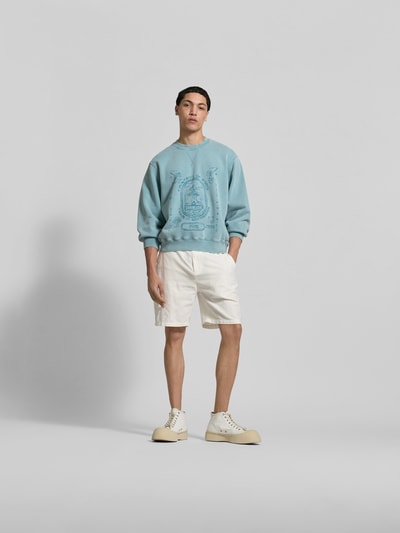 Männermodel trägt Oversized Sweatshirt mit Statement-Print von MAISON KITSUNÉ in Hellblau, elastische Abschlüsse, Used-Look, Ganzkörperansicht von vorne