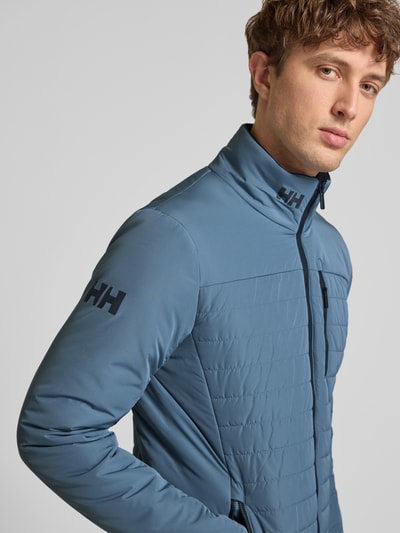 Männermodel trägt Regular Fit Jacke mit Stehkragen von Helly Hansen in blauw met logoprint, steekzakken met ritssluiting, napoleonzak, ruglengte maat M 70 cm, schouderbreedte 46 cm, borstwijdte 54 cm, mouwlengte 69 cm, detailopname bovenlichaam.