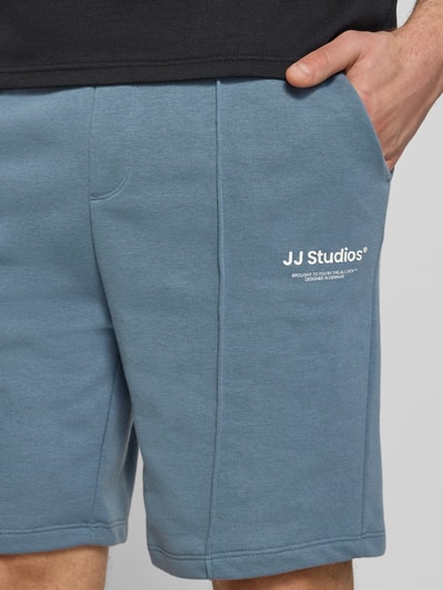 Mężczyzna nosi Sweatshorts mit Logo Model 'KANE' von Jack & Jones z bawełnianego mixu w kolorze niebieskim, krój relaxed fit, z elastycznym pasem, logo i nadrukiem, szerokość w rozmiarze M: 42 cm, długość nogawki od wewnątrz 19 cm, zbliżenie na górną część nóg.