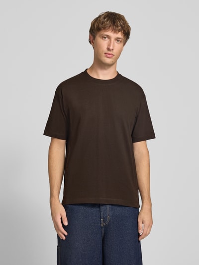Männermodel trägt Relaxed Fit T-Shirt aus reiner Baumwolle von Marc O'Polo DENIM in Dunkelbraun, kombiniert mit blauer Jeans, Ganzkörperansicht von vorne.