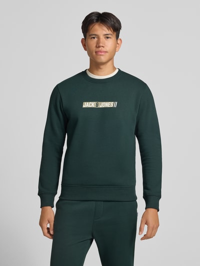 Männermodel trägt Regular Fit Sweatshirt mit Baumwoll-Anteil Modell 'Munk' von JACK & JONES in Dunkelgrün, Logo-Print, Ganzkörperansicht von vorne, Material-Mix, elastische Abschlüsse, Rundhalsausschnitt.
