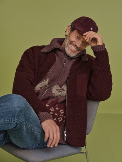 Männermodel trägt Strickjacke mit abstraktem Muster von DRYKORN in dunklem Rot, Baumwolle, gerader Schnitt, Reißverschluss, Nahaufnahme Oberkörper.