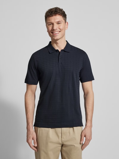 Männermodel trägt Regular Fit Poloshirt aus reiner Baumwolle von Marc O’Polo in Dunkelblau mit Gitterkaro Struktur, Ganzkörperansicht von vorne.