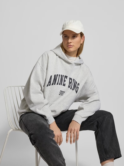 Frauenmodel trägt Oversized Hoodie mit Logo-Print von Anine Bing aus reiner Baumwolle, Oversized-Fit, Känguru-Tasche, elastische Abschlüsse, Rückenlänge 66 cm in Größe S, graue Farbtöne auf glatter Textur, Nahaufnahme Oberkörper.
