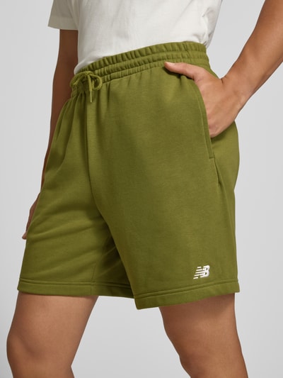 Männermodel trägt Sweatshorts aus Baumwoll-Mix Modell 'FRENCH' von New Balance in Olivgrün mit elastischem Bund und Logo, Nahaufnahme Oberschenkel, Herren Sweatshorts, gerade Passform, Gesäßtasche, Bundweite Größe M 40 cm, Innenbeinlänge 17 cm.