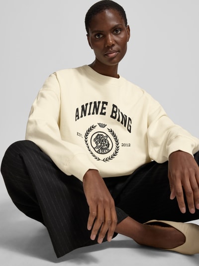 Frauenmodel trägt Oversized Sweatshirt mit Logo-Print von Anine Bing aus reiner Baumwolle, cremefarben mit schwarzem Print, elastische Abschlüsse, Rundhalsausschnitt, Oversized-Fit, Rückenlänge bei Größe S 61 cm, Nahaufnahme Oberkörper.