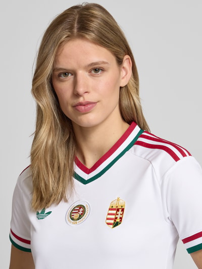 Frauenmodel trägt Ungarn Heimtrikot 2026 von adidas Sportswear in Optik: Unifarben, Regular-Fit, Applikation, Logo-Stitching, Kontraststreifen, V-Ausschnitt, Ärmelform: Kurzarm, Materialverarbeitung: feine Webstruktur, Nahaufnahme Oberkörper.