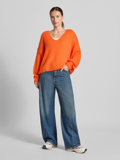 Frauenmodel trägt Oversized Strickpullover mit Mohair-Anteil von Oui, leuchtendes Orange, V-Ausschnitt, grobe Strickstruktur, gerippte Abschlüsse, Kombi mit blauen Jeans, Ganzkörperansicht von vorne.