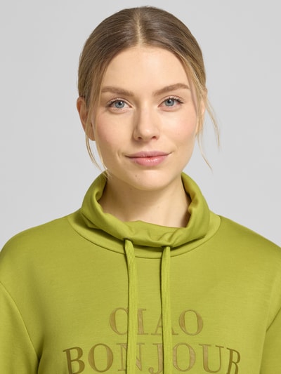 Kobiece model prezentuje Sweatshirt mit Stehkragen und Statement-Print od Soyaconcept, zielony kolor, materiał z domieszką modalu, krój regular fit, nadruk z napisem, stójka z troczkami, zbliżenie na górną część ciała.