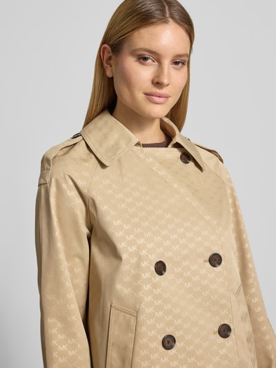 Frauenmodel trägt Trenchcoat mit Logo-Muster von MICHAEL Michael Kors in Beige, Zweireiher, Reverskragen, Ärmelriegel, Nahaufnahme Oberkörper