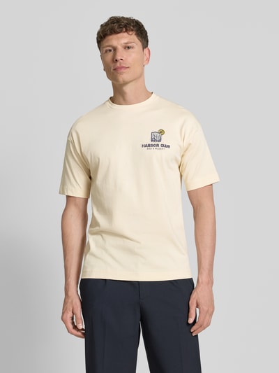 Männermodel trägt T-Shirt mit Motiv-Print und Rundhalsausschnitt von Drykorn in Creme, Reine Baumwolle, Regular-Fit, Rückenlänge 70 cm, Ganzkörperansicht von vorne.