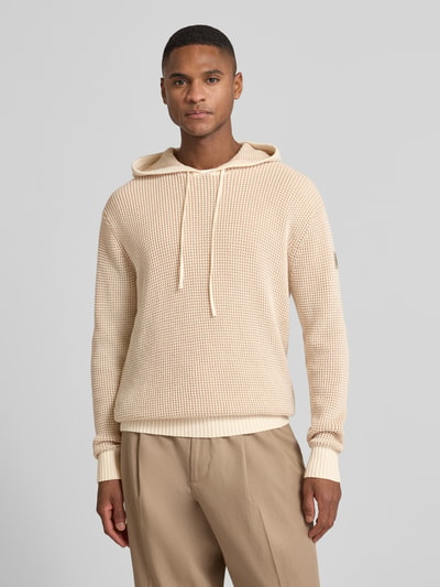 Männermodel trägt Slim Fit Hoodie mit Viskose-Anteil Modell 'Kem' von Guess in Beige, Strick-Optik, elastische Abschlüsse, Ganzkörperansicht von vorne.
