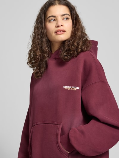 Frauenmodel trägt Hoodie mit Känguru-Tasche von ANOTHER COTTON LAB aus Baumwoll-Mix in Dunkelrot mit Label-Print, Relaxed-Fit, Nahaufnahme Oberkörper.