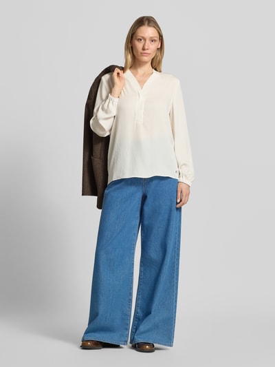 Frauenmodel trägt Bluse mit V-Ausschnitt und Label-Detail von Marc Cain in Creme, kombiniert mit weiter Jeans, elastische Ärmelabschlüsse, Ganzkörperansicht von vorne.