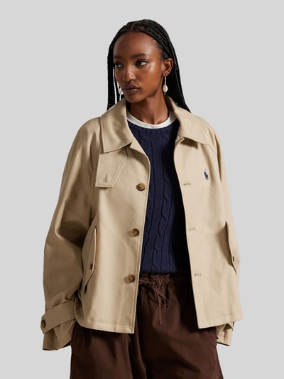 Vrouwenmodel draagt Regular Fit Trenchcoat uit zuivere katoen van Polo Ralph Lauren in uni beige met logo-detail, zijflapzakken, lange mouwen, recht silhouet, eenrijige knopenlijst, kraag met staande optie, detailopname bovenlichaam.