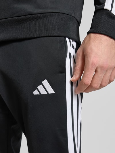 Männermodel trägt Trainingsanzug mit Stretch-Anteil von adidas Sportswear, elastyczna mieszanka materiałów, prosta krój, detale z logo, zamek błyskawiczny, wpuszczane kieszenie, elastyczne wykończenia, stójka, szerokość w rozmiarze M: 39 cm, długość tyłu: 70 cm, szerokość ramion: 44 cm, długość rękawa: 65 cm, długość wewnętrzna nogawki: 73 cm. Detailaufnahme