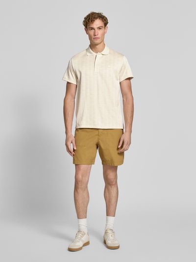 Männermodel trägt Classic Fit Poloshirt mit Logo-Detail von Lacoste in Beige zu ockerfarbenen Shorts, Herren Poloshirt von Lacoste, Ganzkörperansicht von vorne.