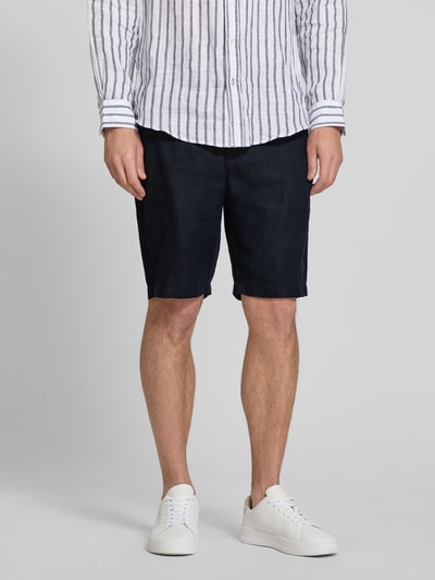 Mężczyzna nosi szorty Leinen Shorts mit Eingrifftaschen Modell 'PANDRUP' od Casual Friday z lnu, krój straight fit, kolor ciemnogranatowy, wpuszczane kieszenie, guziki, zamek błyskawiczny, szerokość w rozmiarze M: 44 cm, długość wewnętrzna nogawki: 23 cm, widok całej sylwetki z przodu.