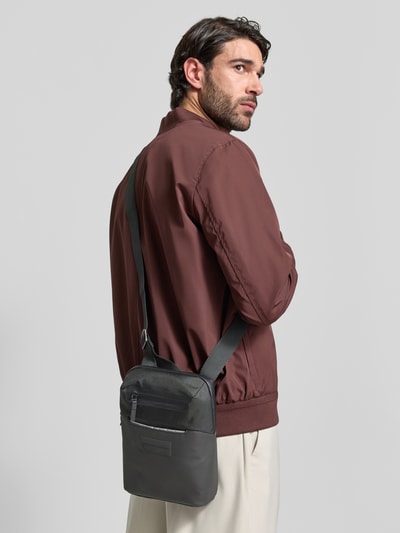 Männermodel trägt Umhängetasche mit Logo-Detail von Porsche Design in Schwarz mit Reißverschlussfach und längenverstellbarem Schulterriemen, Szerokość w rozmiarze 20 cm, długość 26 cm, głębokość 6 cm, długość paska 68 cm, Ganzkörperansicht Rückansicht