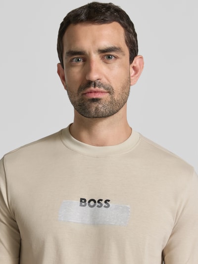 Männermodel trägt Regular Fit T-Shirt aus reiner Baumwolle Modell 'Te_LOGOTAPE' von BOSS Orange in Beige, Motiv-Print vorne, Nahaufnahme Oberkörper.