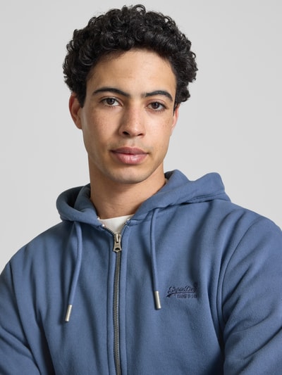 Männermodel trägt Sweatjacke mit Kapuze und Logo-Stitching von Superdry in Blau, weiche Textur, Reißverschluss, Nahaufnahme Oberkörper. Baumwoll-Mix, Regular Fit, Kapuze mit Kordelzug, Logo-Stitching, seitliche Eingrifftaschen, gerippte Abschlüsse, Label-Detail.