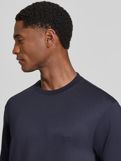 Männermodel trägt T-Shirt mit Logo-Stitching von Emporio Armani in Dunkelblau, glatte Textur, Rundhalsausschnitt, Nahaufnahme Oberkörper. Elastischer Modal-Mix mit Seiden-Anteil, gerade Passform, Rückenlänge bei Größe M 72 cm.
