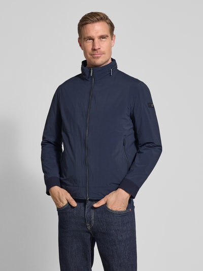 Mężczyznamodel nosi Regular Fit Jacke mit Stehkragen od Bugatti, granatowy kolor, zamek błyskawiczny dwustronny, dwie wpuszczane kieszenie z zamkiem błyskawicznym, detal z logo, kaptur chowany, zakończenia rękawów elastyczne, długość tyłu w rozmiarze 50: 65 cm, szerokość ramion w rozmiarze 50: 47 cm, szerokość klatki piersiowej w rozmiarze 50: 53 cm, długość rękawów w rozmiarze 50: 63 cm, całopostaciowy widok z przodu.