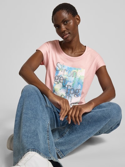Frauenmodel trägt T-Shirt mit Statement-Print von Christian Berg Woman in Rosa, Baumwoll-Viskose-Mix, Regular-Fit, Rundhalsausschnitt, Rückenlänge bei Größe S: 57 cm, kombiniert mit blauen Jeans, sanfte Farben, glatte Textur, Nahaufnahme Oberkörper.