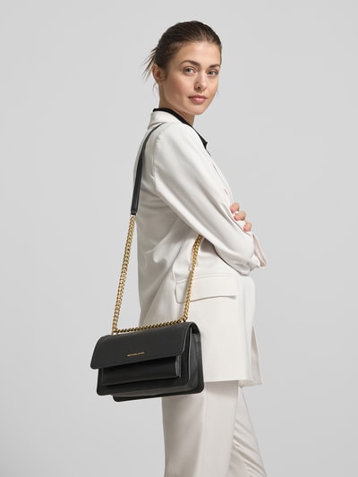 Frauenmodel trägt Umhängetasche aus Leder mit Logo-Detail Modell 'CLAIRE' von MICHAEL Michael Kors, schwarze Farbe, goldfarbener Schultergurt in Gliederketten-Optik, glatte Textur, Hauptfach mit Umschlag und Druckknopfverschluss, Reißverschlussfach innen, Größe 23x15x10 cm, Länge Schulterriemen 110 cm, Ganzkörperansicht von vorne.