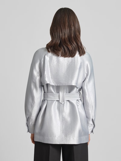 Frauenmodel trägt Jacke aus Viskose-Mix mit Reverskragen und Gürtel von Marc Cain in silber, glatte Oberfläche, Ganzkörperansicht Rückansicht. Taillierter Schnitt, Knopfverschluss, seitliche Eingrifftaschen, Maße bei Größe 36.