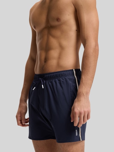 Männermodel trägt Badeshorts mit Tunnelzug Modell 'ICONIC' von BOSS, dunkelblau, label print, kontraststreifen, elastischer bund, seitentaschen, achterzak. Ganzkörperansicht von vorne.