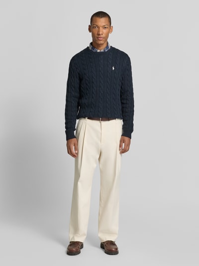 Männermodel trägt Pullover mit Logo-Stitching von POLO RALPH LAUREN in Dunkelblau mit Zopfmuster, cremefarbene Hose, Ganzkörperansicht von vorne.