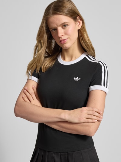Kobiece modelka nosi Slim Fit T-Shirt aus Baumwoll-Mix od adidas Originals, efekt: jednolity kolor, krój regular fit, wyhaftowane logo, paski w kontrastowym kolorze, okrągły dekolt, krótki rękaw, materiał: mieszanka bawełny. Ujęcie: z przodu, górna część ciała.