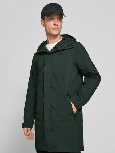 Männermodel trägt Regular Fit Mantel mit Kapuze Modell 'Brussel' von Helly Hansen in Dunkelgrün, glatte Textur, Ganzkörperansicht von vorne. Rückenlänge 96 cm, Schulterbreite 46 cm, Brustweite 51 cm, Ärmellänge 66 cm, Logo-Detail.