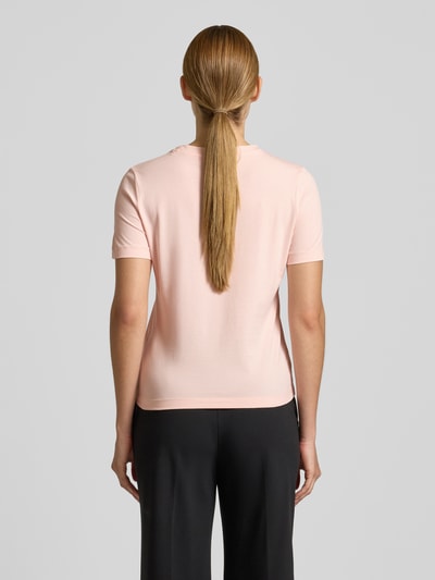 Frauenmodel trägt T-Shirt mit Strasssteinbesatz Modell 'SIENA' von Guess Activewear in zartem Rosa, glatte Baumwollstruktur, Ganzkörperansicht Rückansicht.