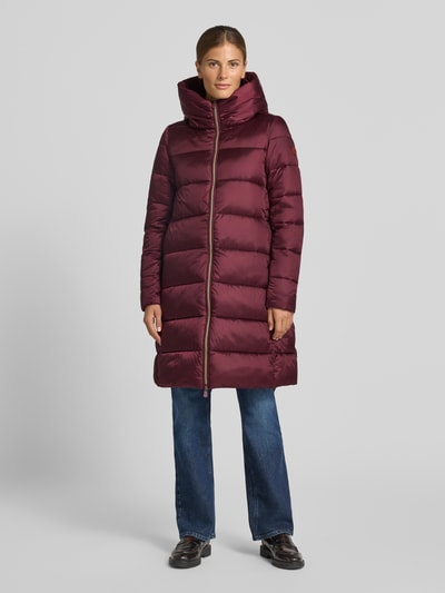 Frauenmodel trägt bordeauxfarbenen Steppmantel mit Kapuze Modell 'LYSA' von Save The Duck, gerader Schnitt, Ganzkörperansicht von vorne.