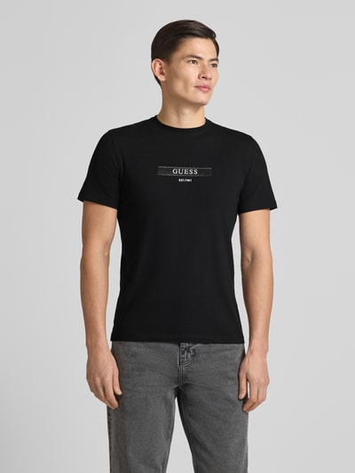 Männermodel trägt T-Shirt mit Label-Detail von Guess in Schwarz, Rundhalsausschnitt, Regular Fit, schlichte Optik, Ganzkörperansicht von vorne.