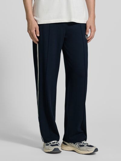 Männermodel trägt trainingsbroek uit mix van viscose en elastaan van EA7 Emporio Armani met recht pasvorm, galonstrepen, labelprint, steekzakken, elastische band, donkerblauw met witte details. Ganzkörperansicht van voren.