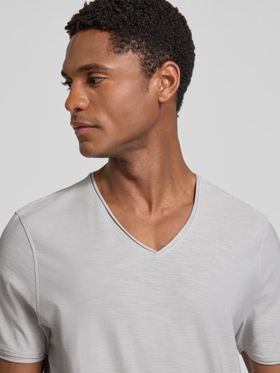 Męski model nosi T-Shirt mit V-Ausschnitt und Rollsaum od MCNEAL z bawełny, krój regular fit, dekolt w serek, jasnoszary, zrolowany brzeg, długość tyłu w rozmiarze M: 81 cm, zbliżenie na górną część ciała.