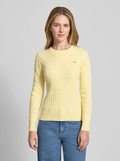 Frauenmodel trägt Strickpullover mit Zopfmuster und Label-Stitching von Gant, gelb, Baumwoll-Mix mit Stretch-Anteil, Slim Fit, Rundhalsausschnitt, Rückenlänge 58 cm, Ärmellänge 61 cm, Ganzkörperansicht von vorne.