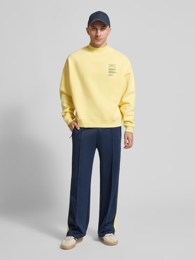 Männermodel trägt Sweatshirt mit Statement-Prints Modell 'Athletic Department' von ANOTHER COTTON LAB in Gelb, Baumwoll-Mix, Loose Fit, kombiniert mit blauer Hose, Ganzkörperansicht von vorne.
