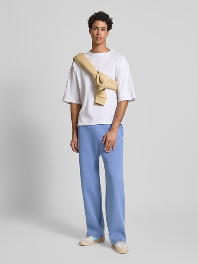 Männermodel trägt Loose Fit Sweatpants mit Seitentaschen von REVIEW aus Baumwoll-Mix in Blau mit Label-Print, Ganzkörperansicht von vorne.