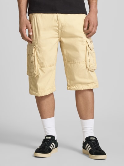 Mannenmodel draagt Bermudas met Pattentaschen en Logo-Stitching van Alpha Industries in beige, straight fit katoen met klep- en steekzakken, knoop- en ritssluiting, vooraanzicht volledige lichaamsopname.