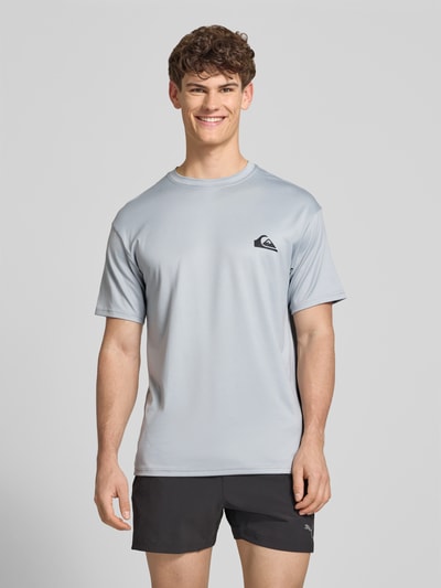 Männermodel trägt T-Shirt mit Rundhalsausschnitt und Logo von Quiksilver in Hellgrau, Regular Fit, glatte Textur, Ganzkörperansicht von vorne